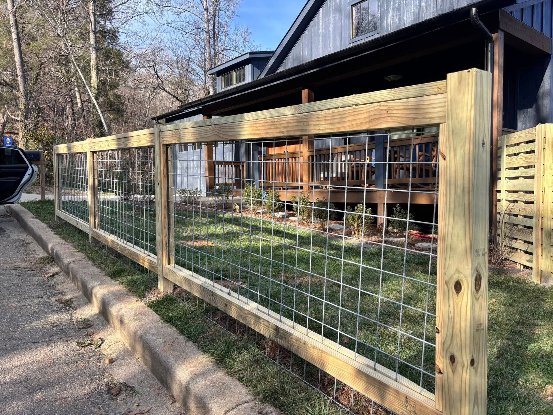 A wooden post-and-rail fence with wire grid paneling along a gravel path in front of a house.