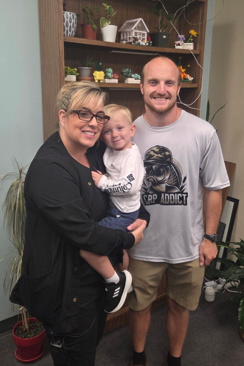 Woman holding child, stands next to man in front of shelf with plants and decor.