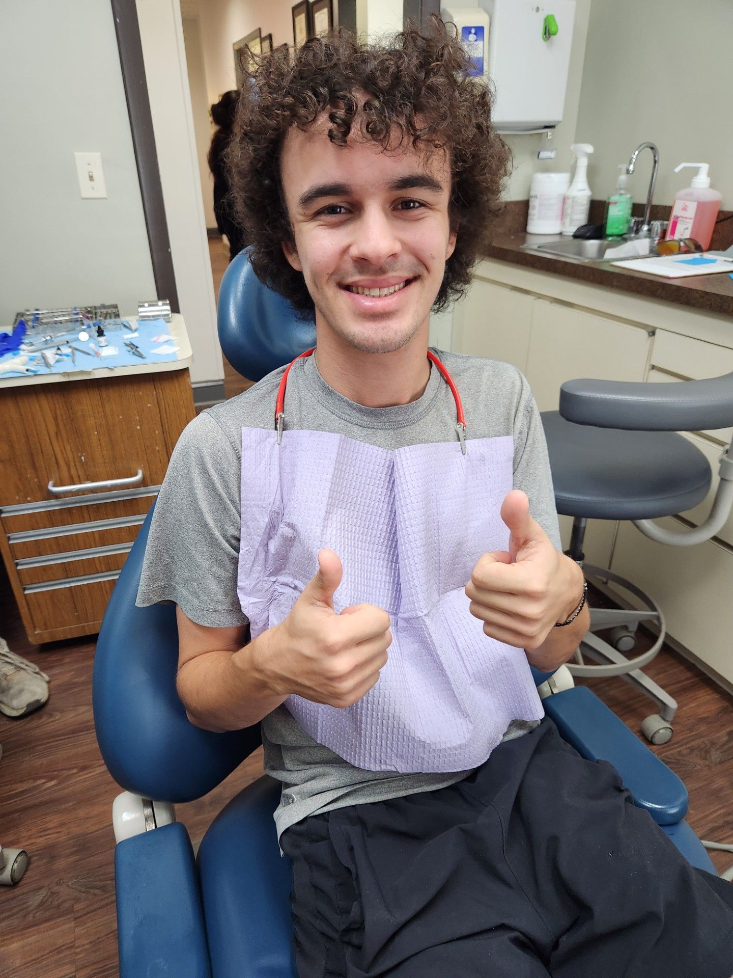 Young man in a dental chair gives thumbs up, smiling; wearing bib. Brown hair.