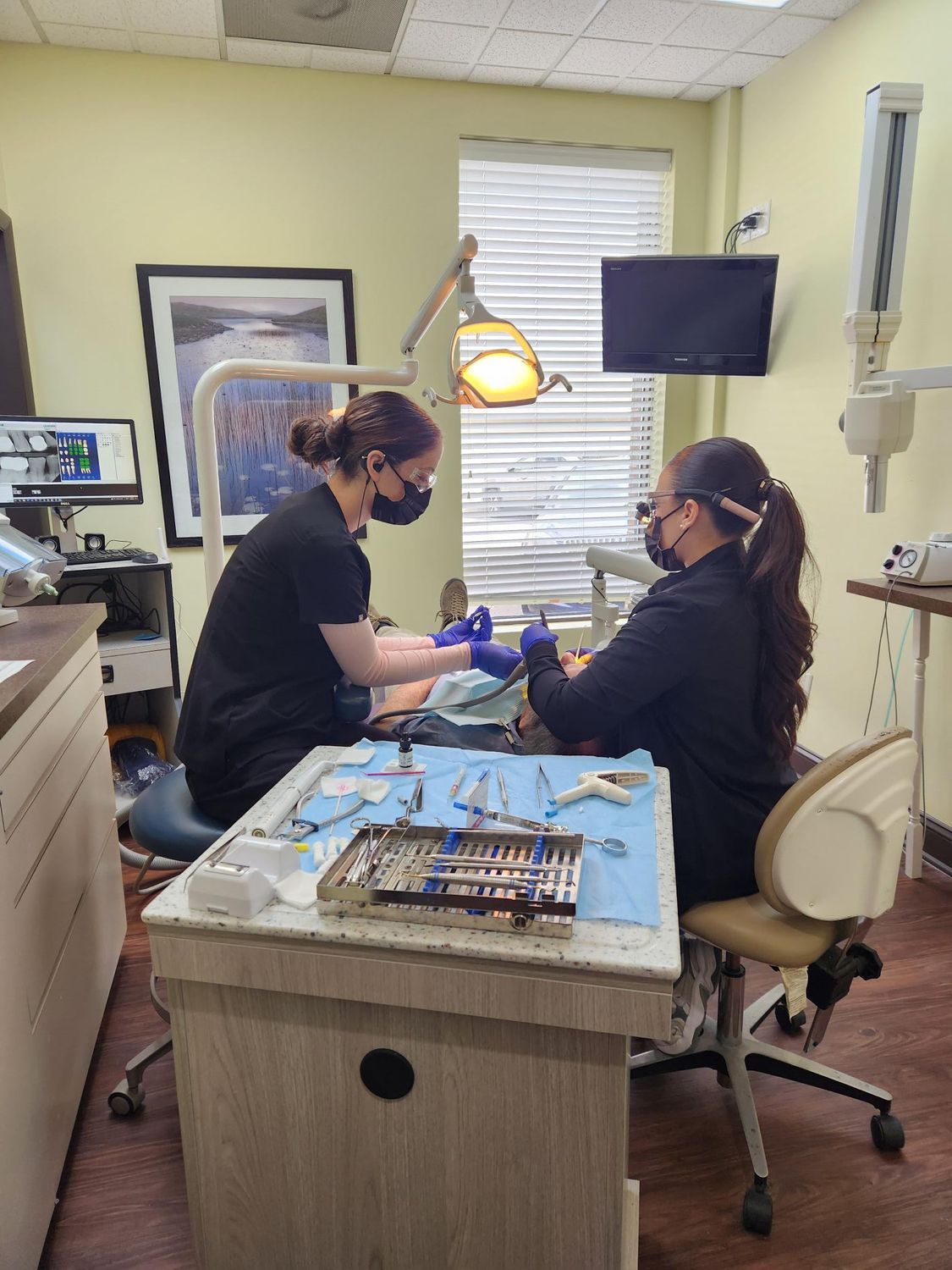 Two dental professionals in black scrubs at work; tools on the table, the patient in the chair.