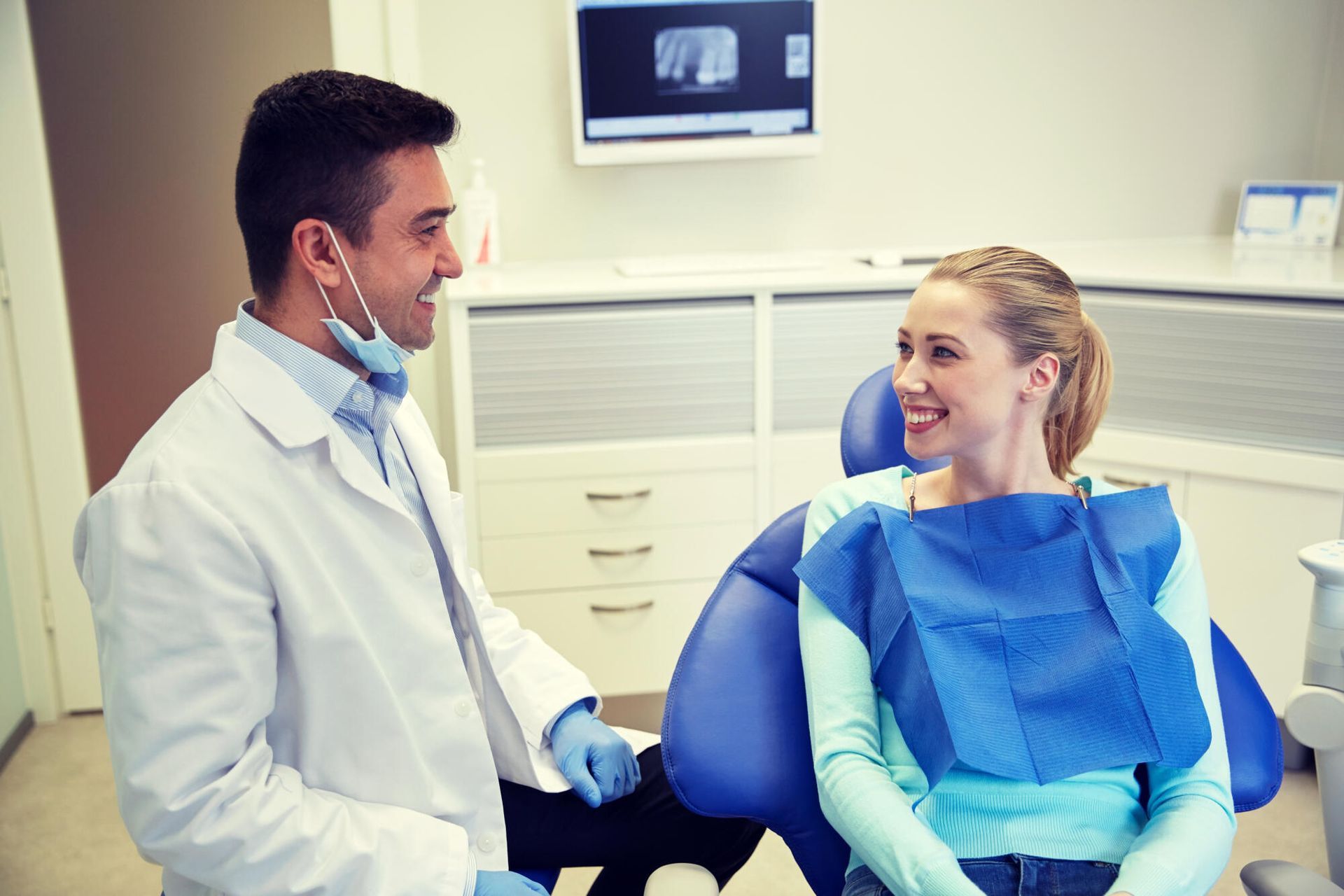 Dentist in a white coat smiles at a woman in a dental chair, a blue paper bib around her neck.