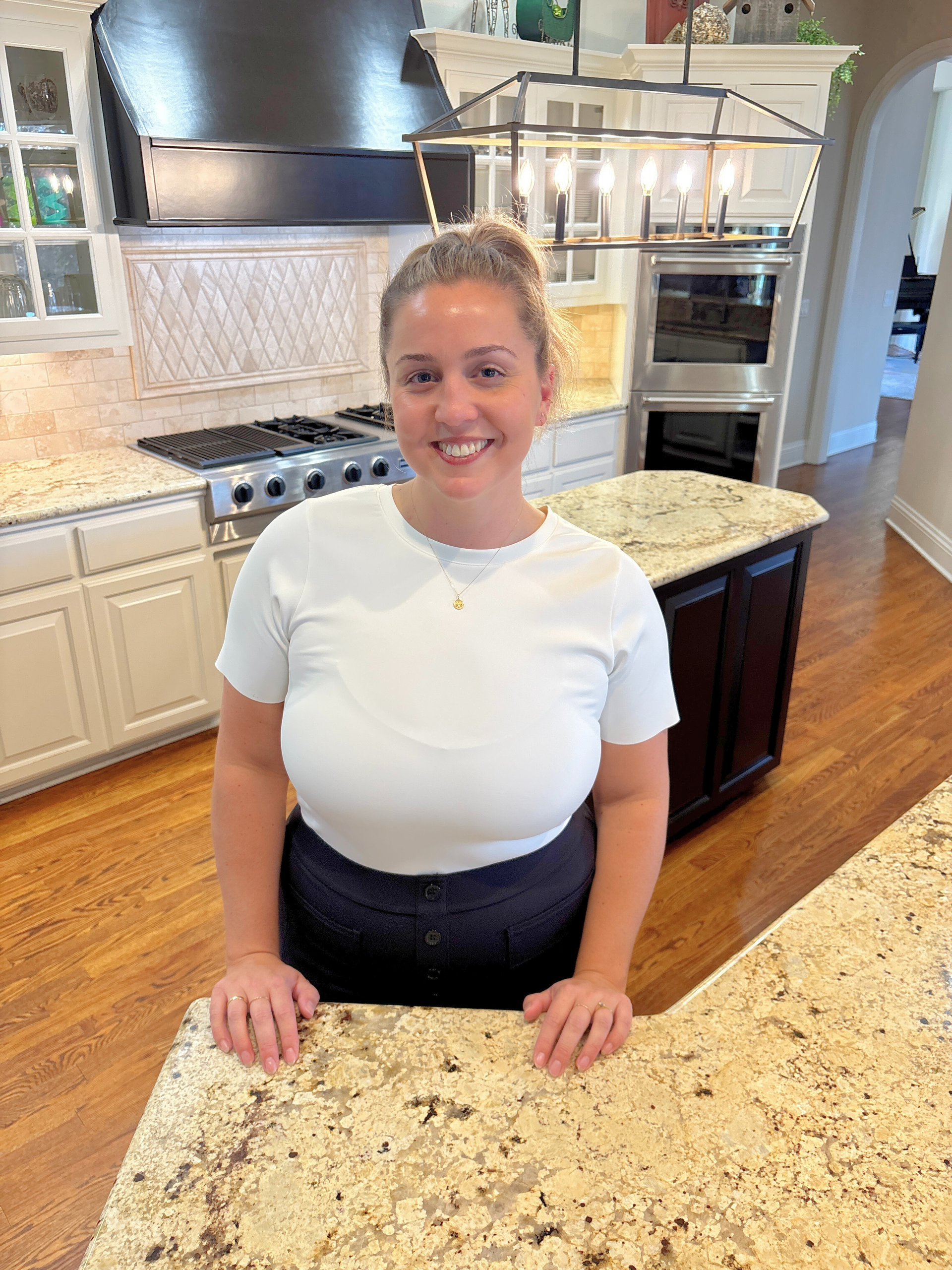 Woman in white shirt smiles in a kitchen, leaning on a counter.