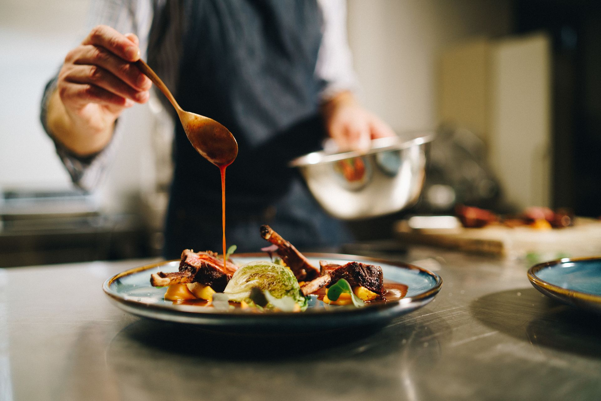 Chef plating food, drizzling sauce onto a plate with meat, vegetables. Kitchen setting.