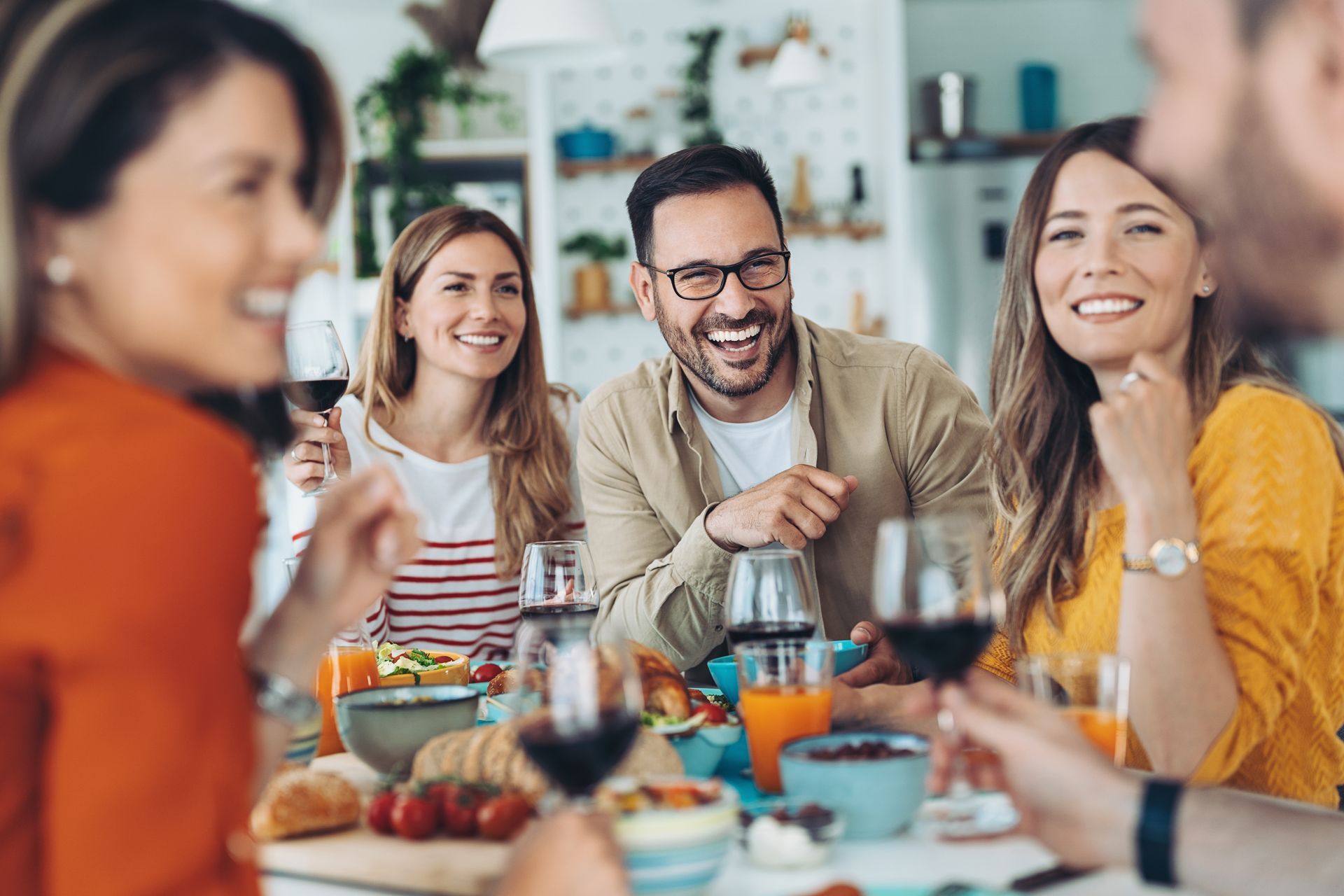 People smiling, drinking wine, and eating at a table, likely friends or family, in a brightly lit kitchen.