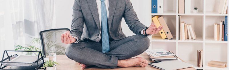 A man in a suit and tie is sitting on a desk in a lotus position.