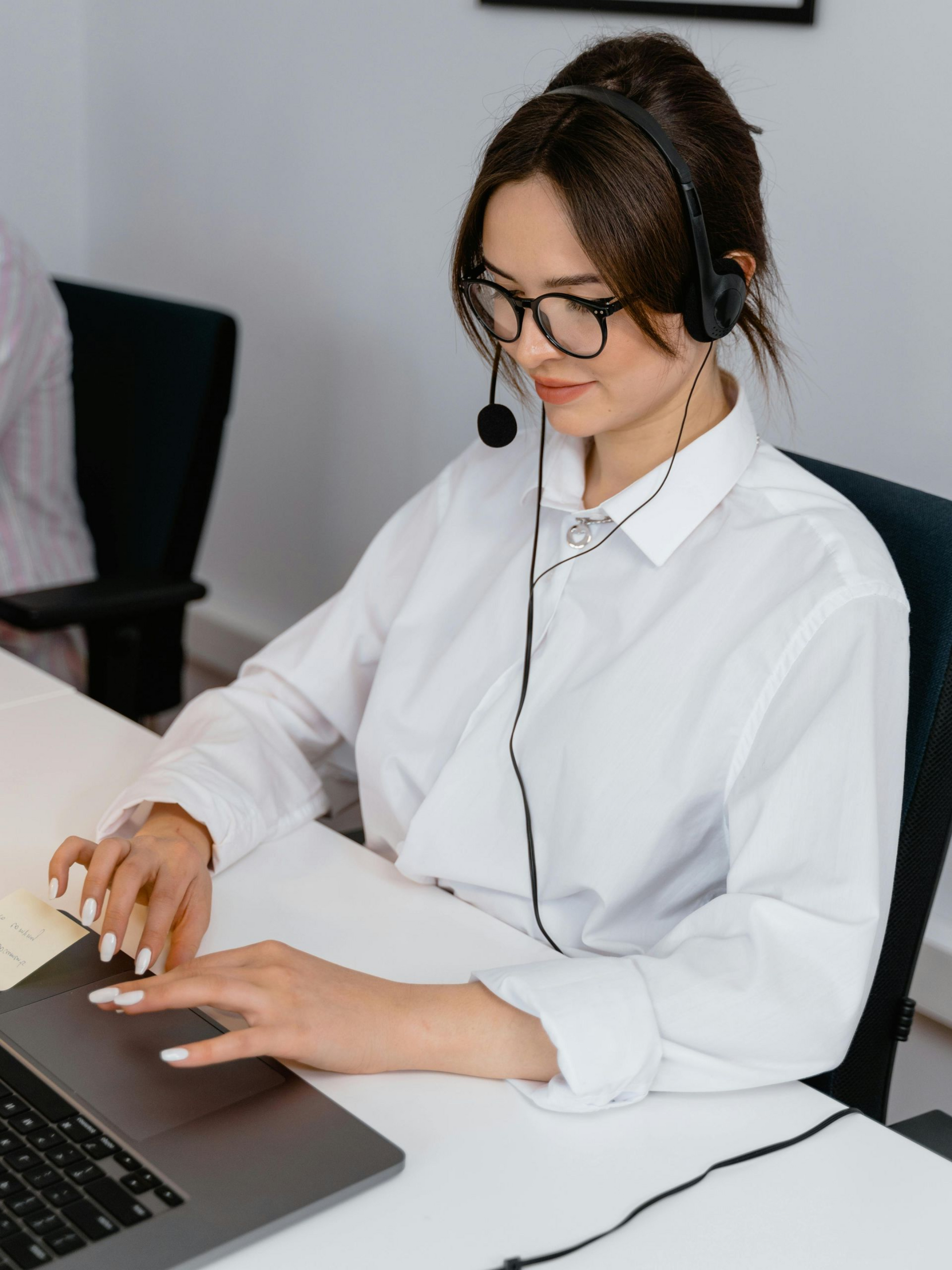 Woman wearing headset and glasses, working on a laptop at a desk.