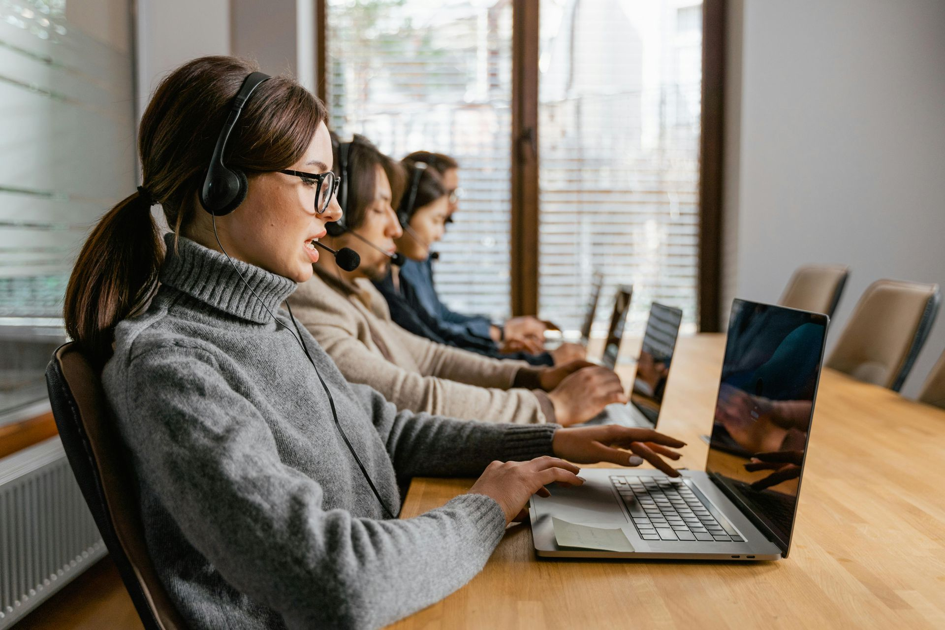 People wearing headsets work on laptops at a long wooden table in a well-lit office.