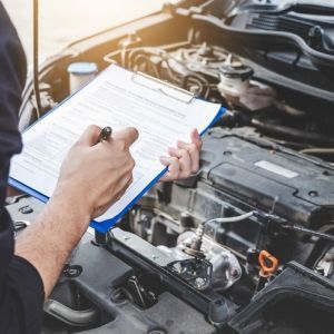 A man is writing on a clipboard in front of a car engine.