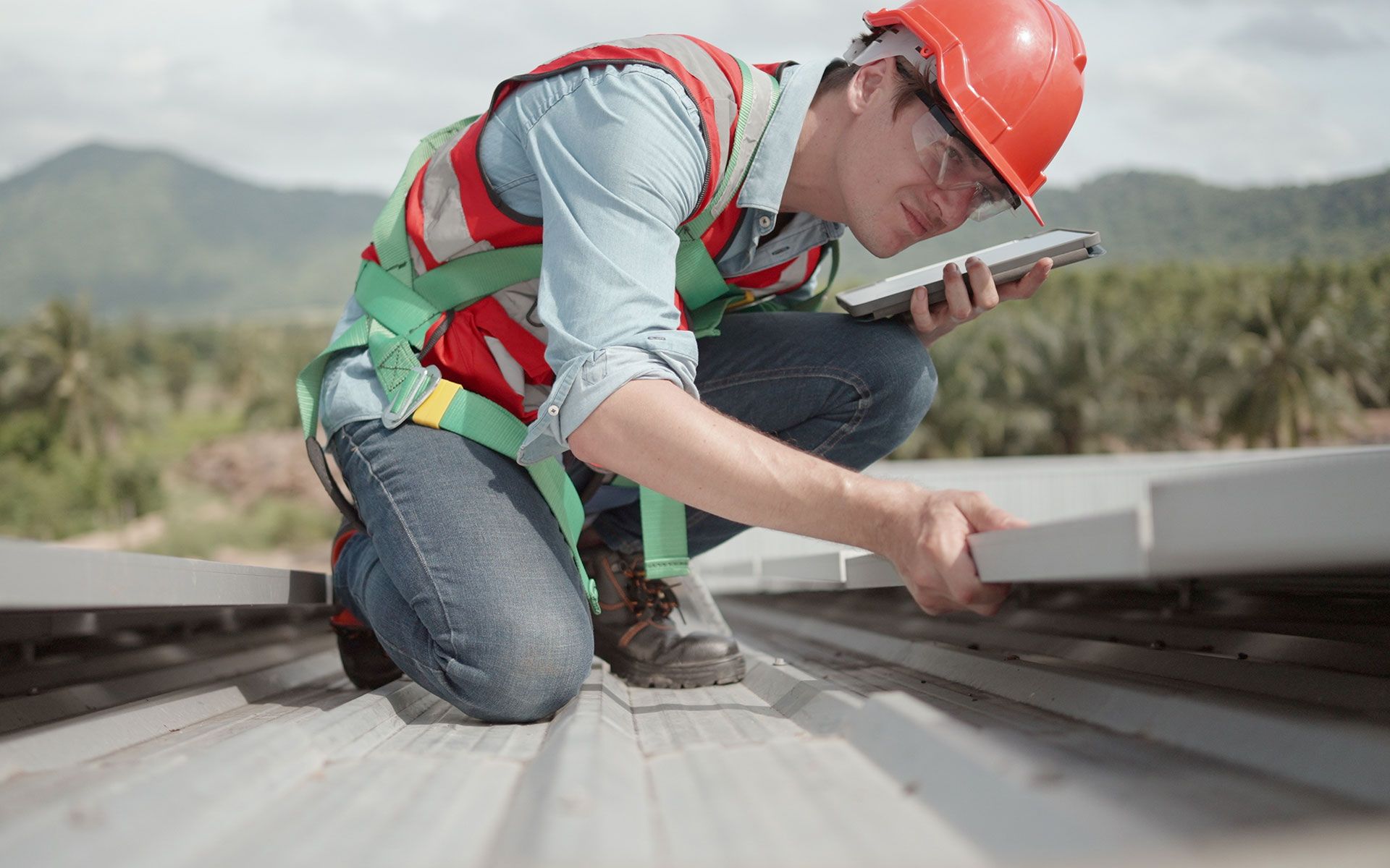 A construction worker is kneeling down on a roof and looking at a tablet.