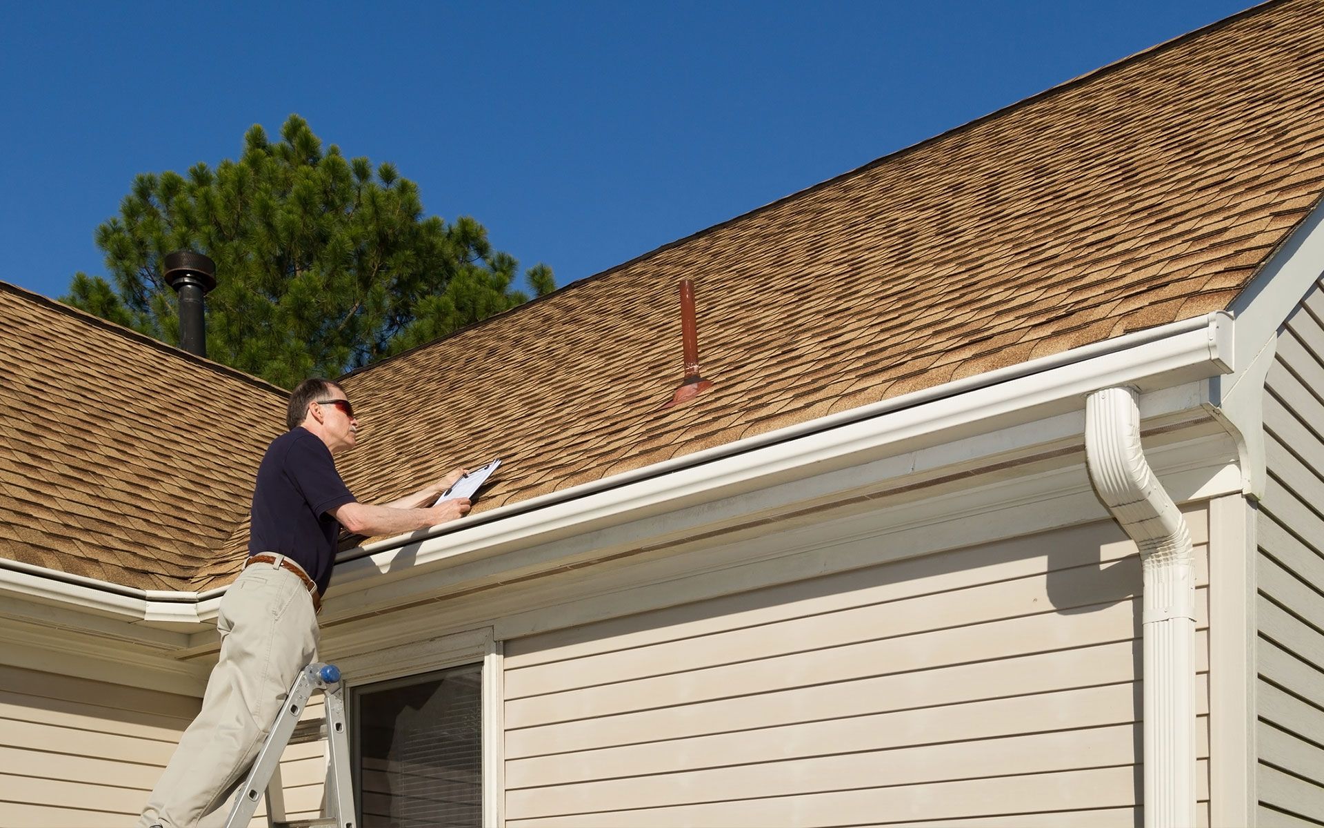 A man is standing on a ladder looking at the roof of a house.