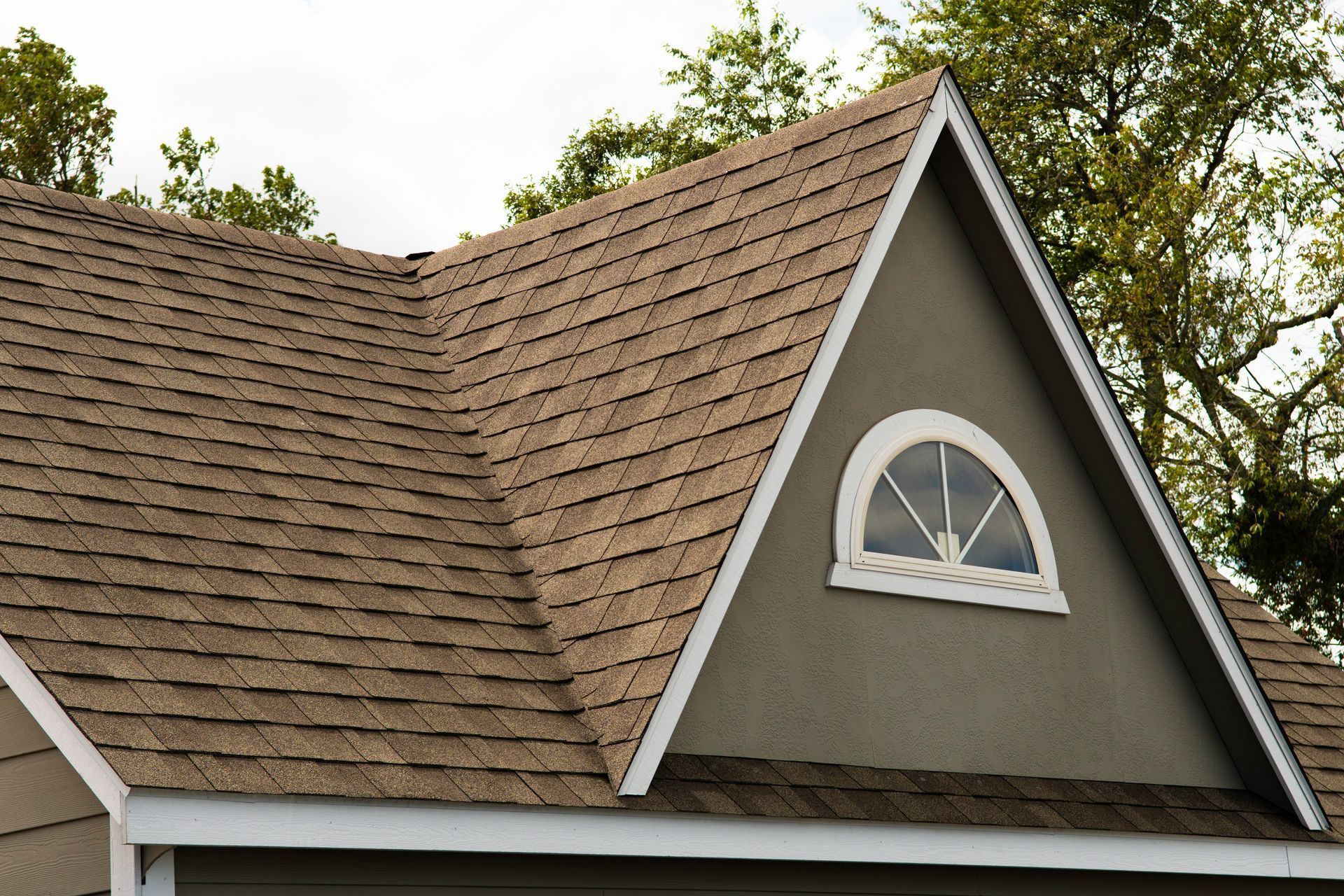 House roof with brown shingles and a small arched window under trees