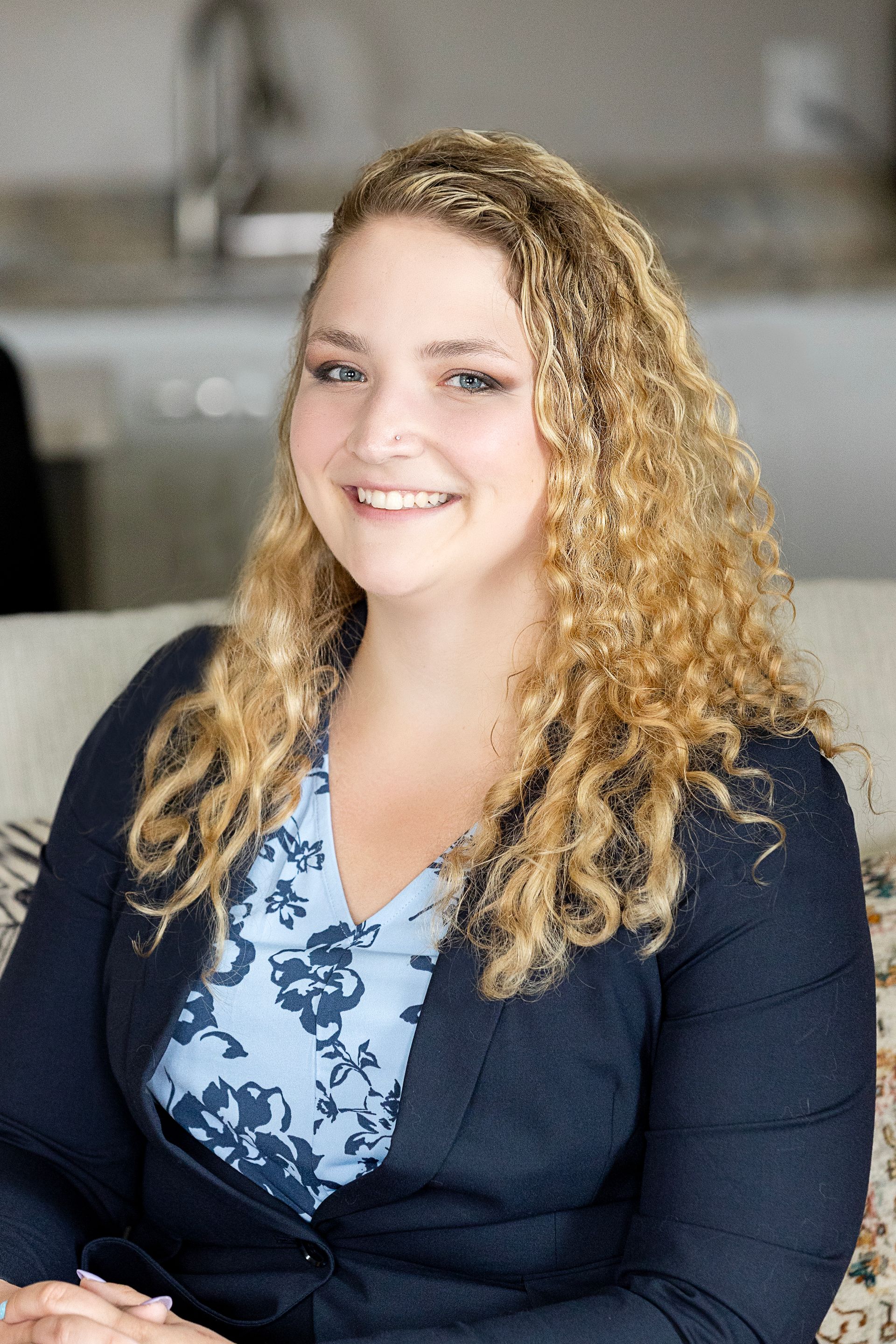 A woman with curly hair is sitting on a couch and smiling for the camera.