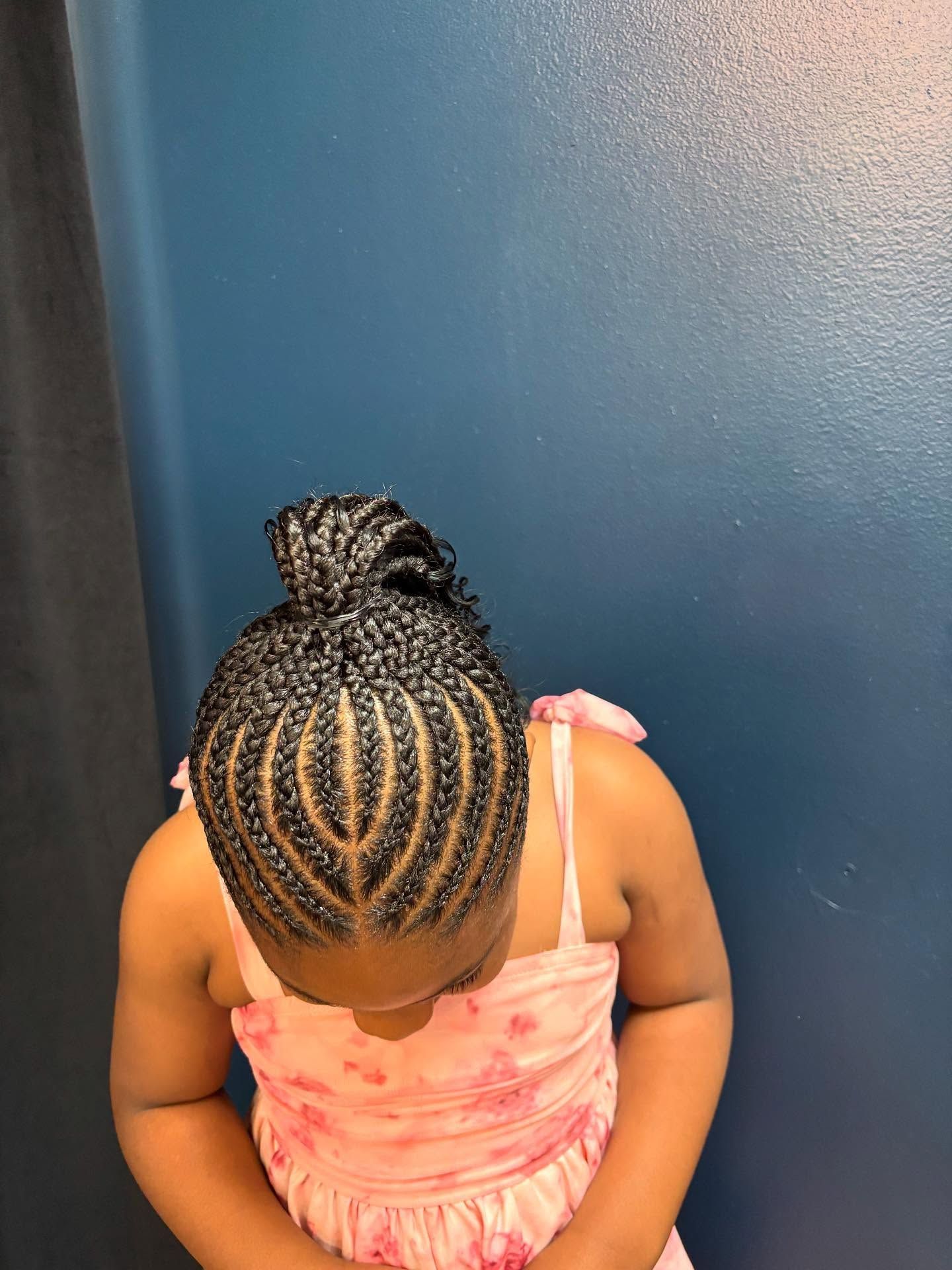 Girl with braided hairstyle, wearing pink dress, looking down against a blue wall.