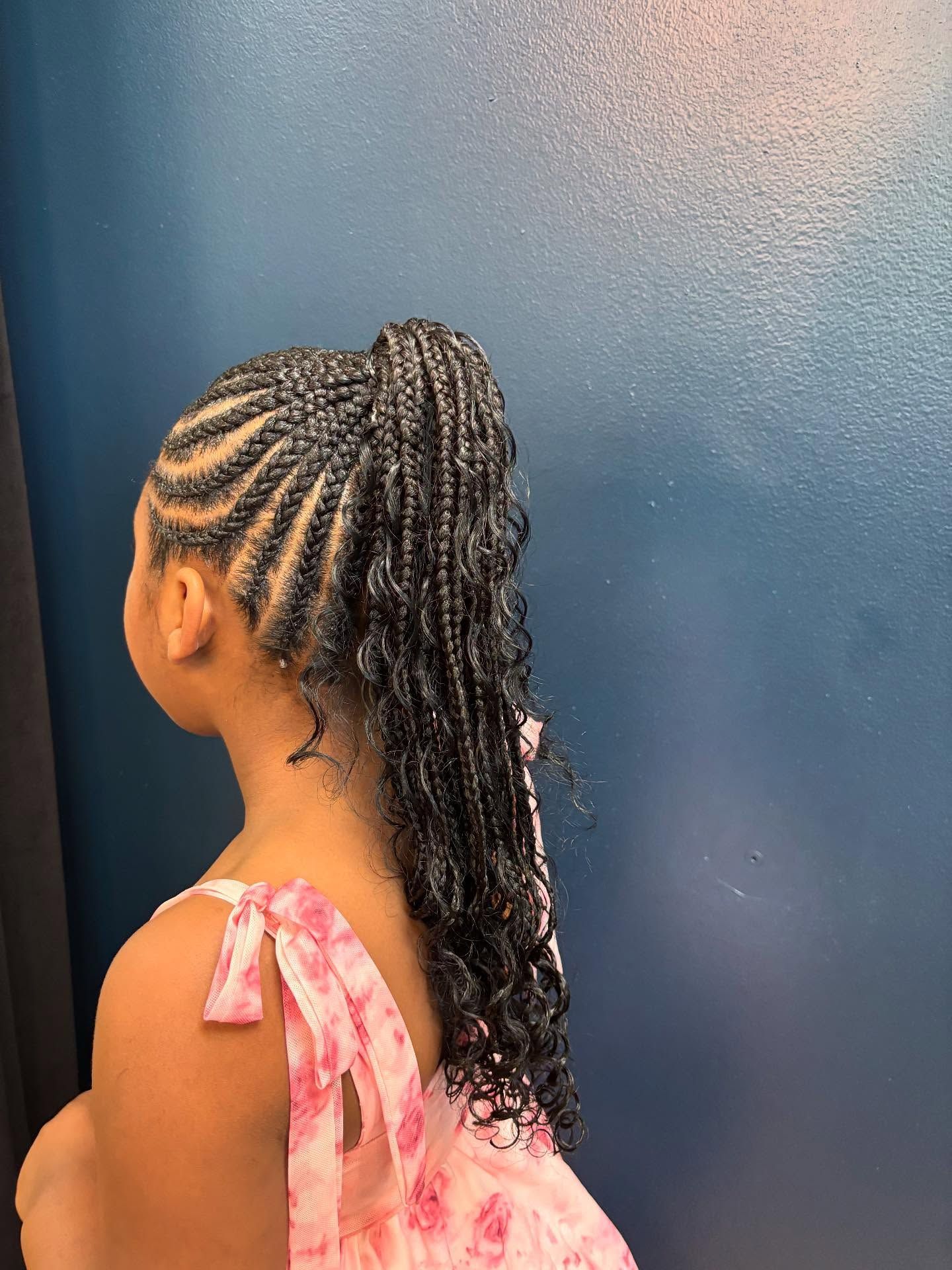 A child with black braids in a high ponytail against a teal wall, wearing a pink floral dress.
