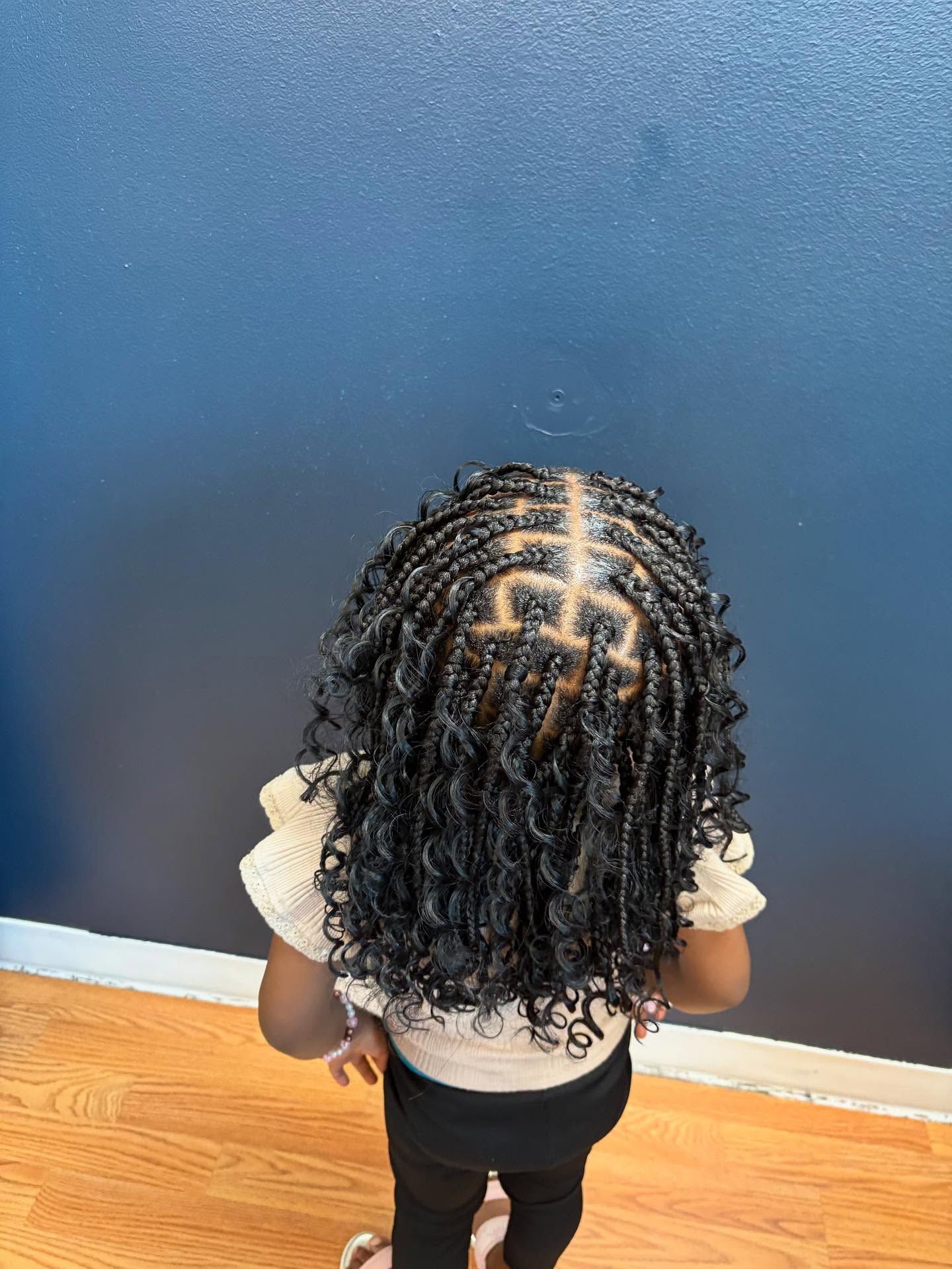 Child with small braids, center part design, black hair, wearing a white top and black pants, standing against a blue wall.