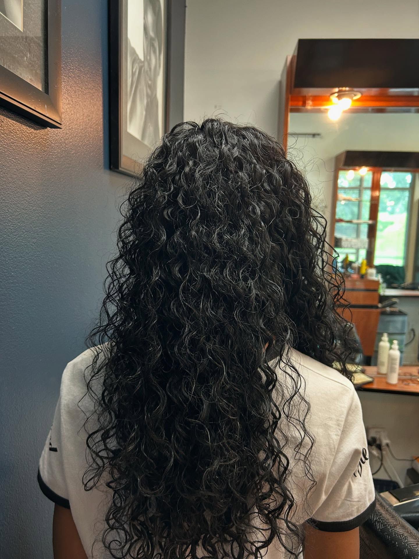 Woman with long, dark, curly hair; standing in a salon.