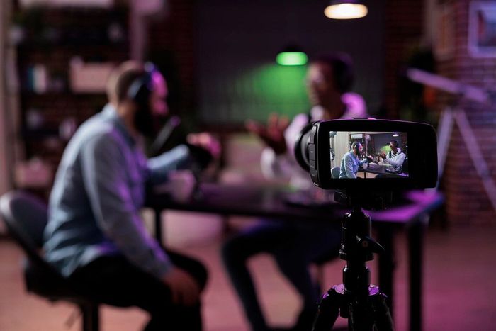 A man is sitting at a table talking to another man in front of a camera.