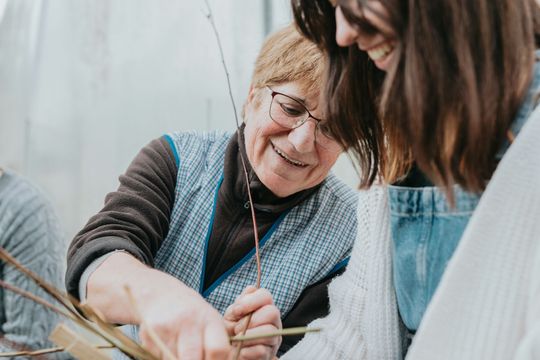 Woman in glasses smiles, shows craft to another smiling person. Indoor, close-up.