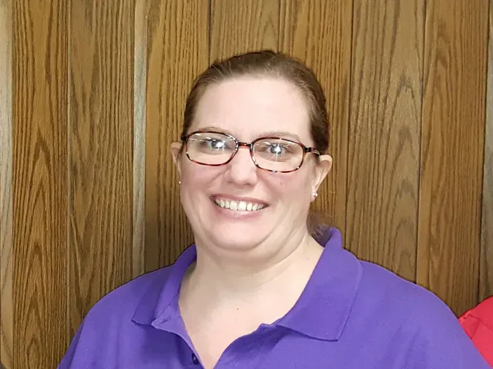 Woman in purple shirt and glasses smiles at the camera, wooden panel background.