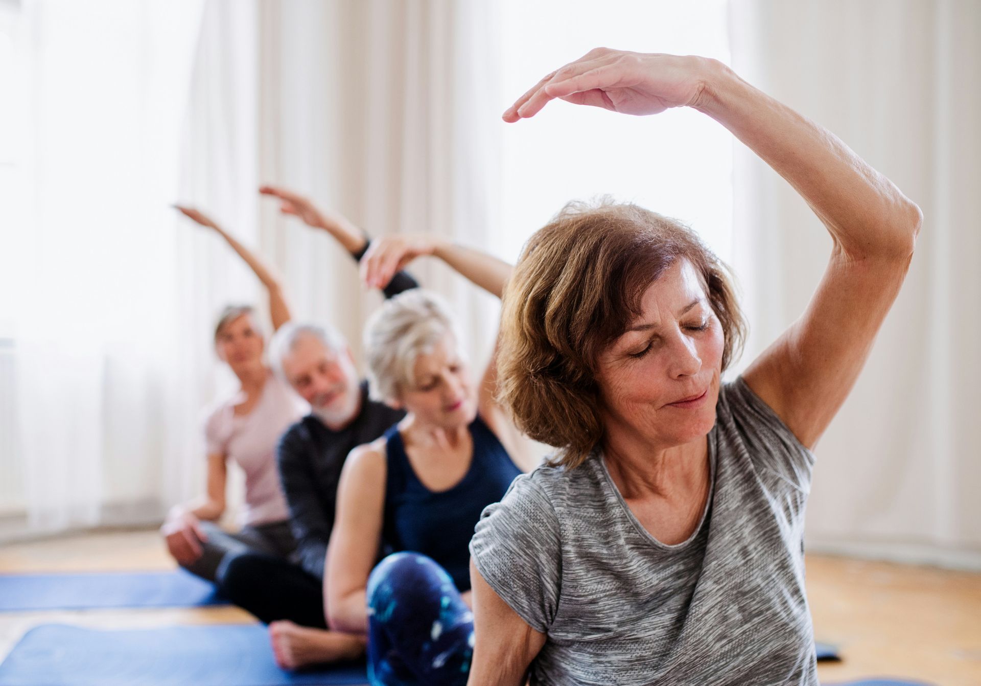 People in a row doing seated side stretch in a brightly lit room, arms raised.