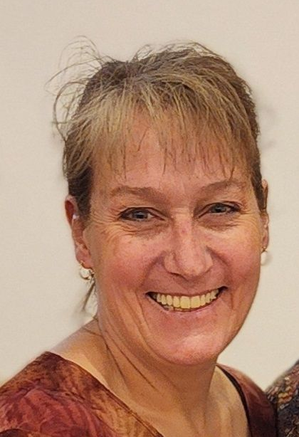 Woman with short, light brown hair smiles broadly, wearing a colorful patterned top; neutral background.