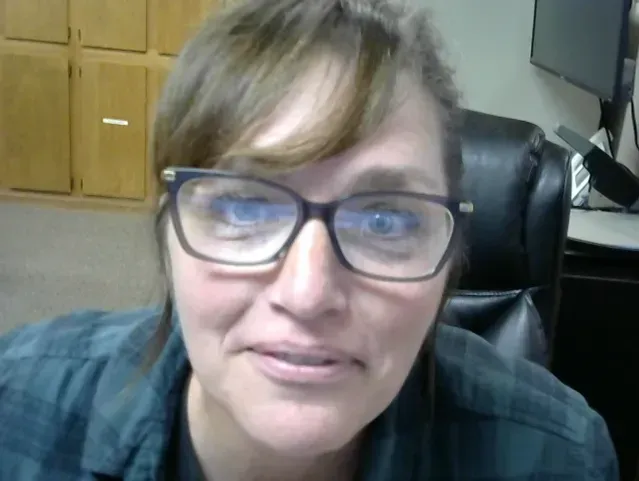 Woman with glasses, looking at the camera. Wearing a dark plaid shirt. Sitting in an office.