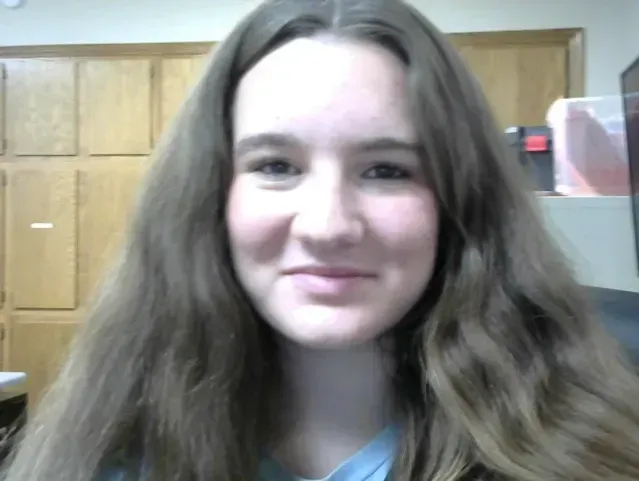 Young person with long brown hair smiling in front of wooden cabinets.