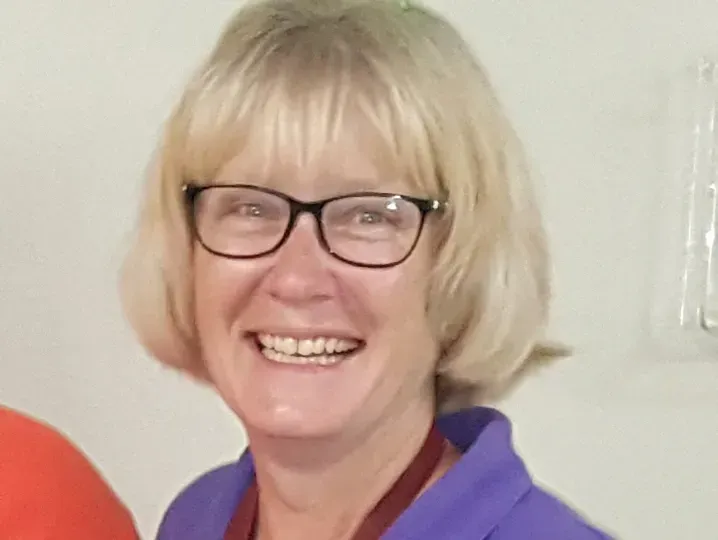 Woman with blonde hair, glasses, smiling, wearing a purple shirt, indoors.