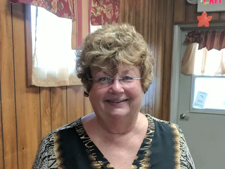 Woman with glasses and short, curly hair smiling indoors, wearing a patterned top.