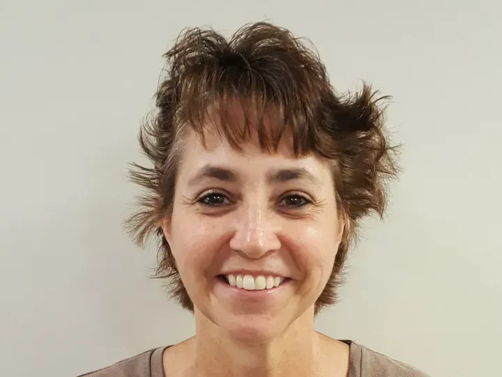 Woman with short, spiky brown hair smiles, facing the camera. Light skin, neutral background.