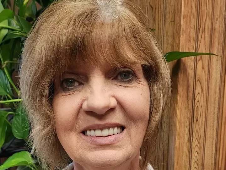 Woman with light brown hair smiles in front of wood paneling and green plants.