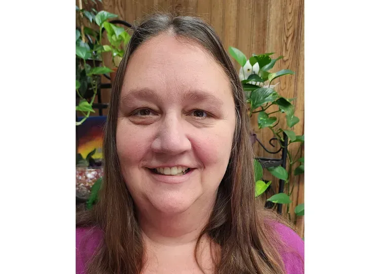 Woman with long brown hair smiles, purple shirt, background with plants.