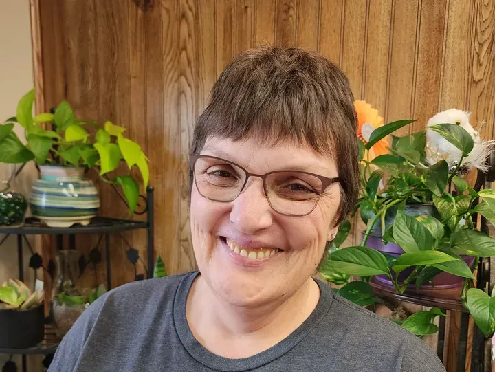 Woman smiling, wearing glasses and a gray shirt, with houseplants in the background.