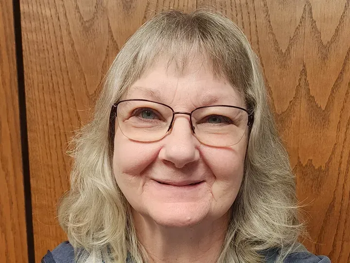 Woman with glasses, gray hair, smiling, in front of a wood-paneled wall.