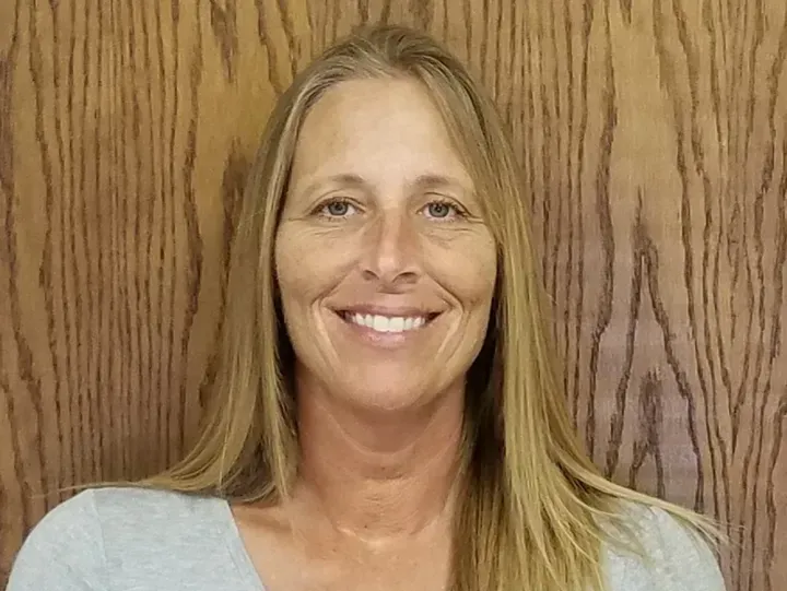 Woman with blonde hair smiling, wearing a gray shirt, in front of a wood-grain wall.