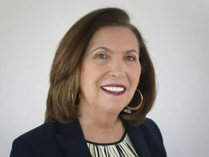 Woman with dark hair smiles, wearing a navy blazer and gold hoop earrings.