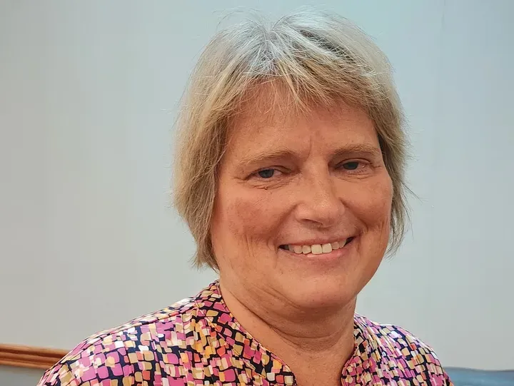 Woman smiling, light hair, colorful patterned shirt, against a pale blue background.