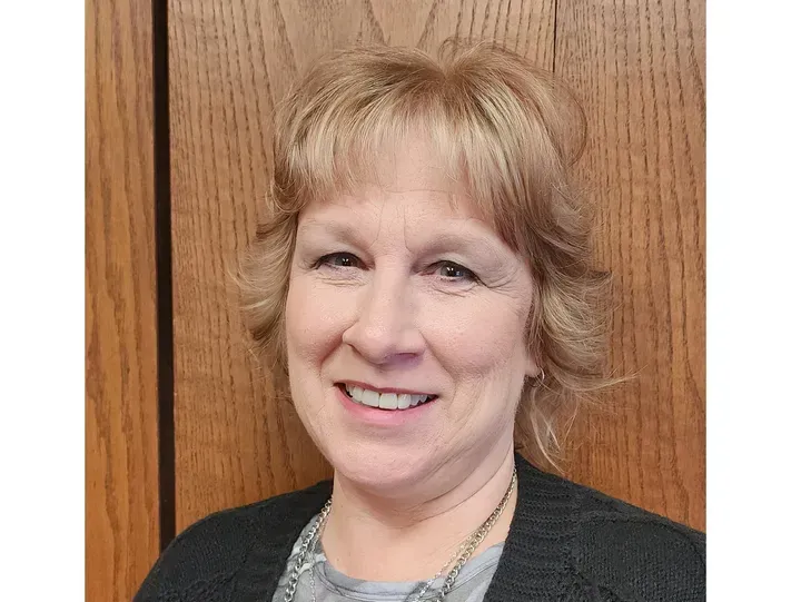Woman with short, layered blonde hair smiles at the camera. She wears a dark sweater. Wooden background.