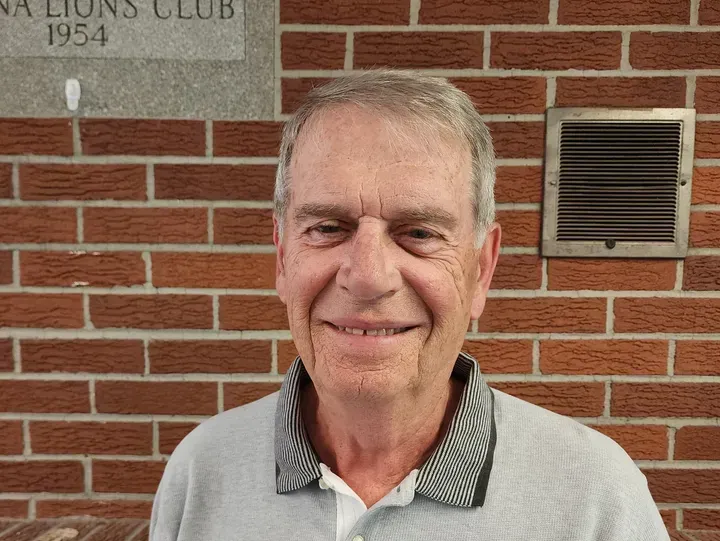 Smiling man in a light blue shirt, standing in front of a brick wall and a stone sign for a Lions Club.