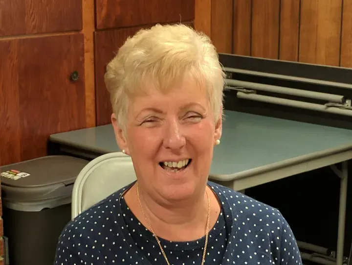 Woman with blonde, short hair smiles widely; wearing a blue polka-dotted shirt; indoors, with a table and wooden paneling.
