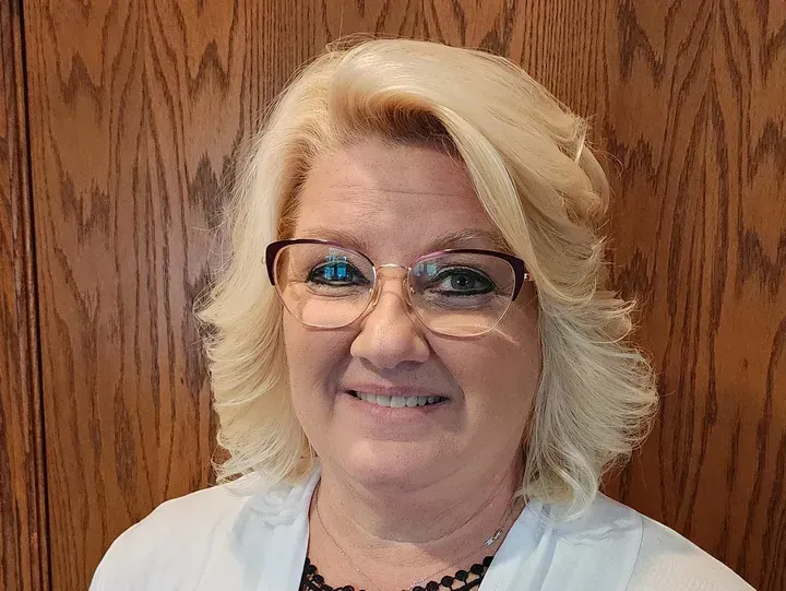 Woman with blonde hair and glasses smiles in front of a wood-paneled wall.