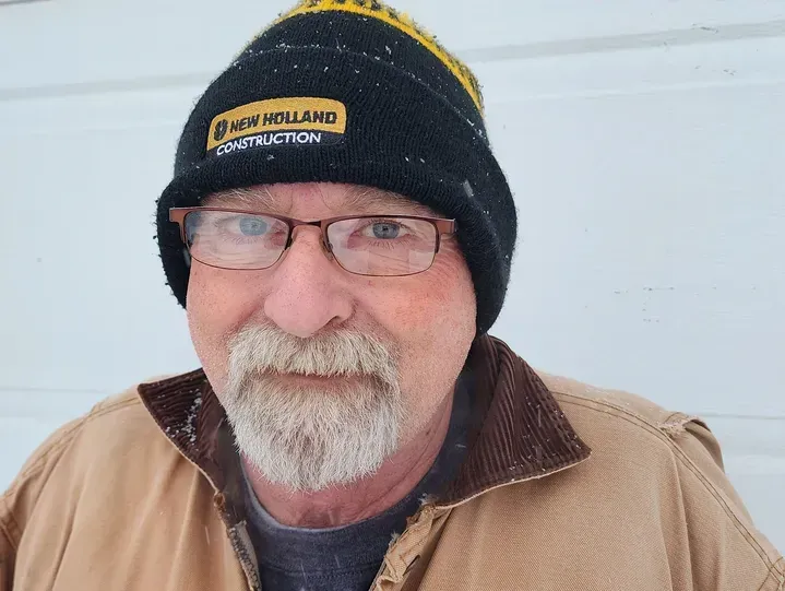 Man wearing a black hat, glasses, and a brown coat, outside in front of a white surface.