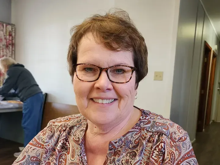 Woman smiling, wearing glasses and patterned top, seated indoors. Another person works in the background.