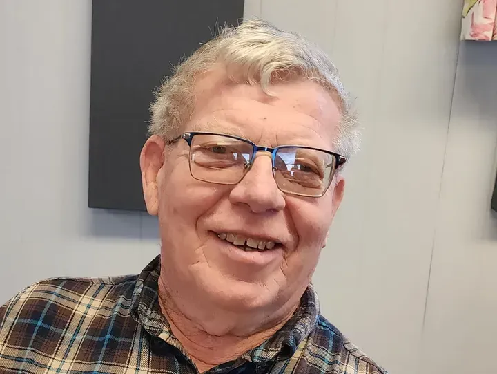 Man wearing glasses smiles, plaid shirt. Indoor setting with gray and white walls.