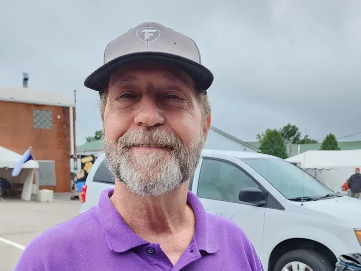Man with a gray cap and purple shirt smiles, standing outdoors near a white van.