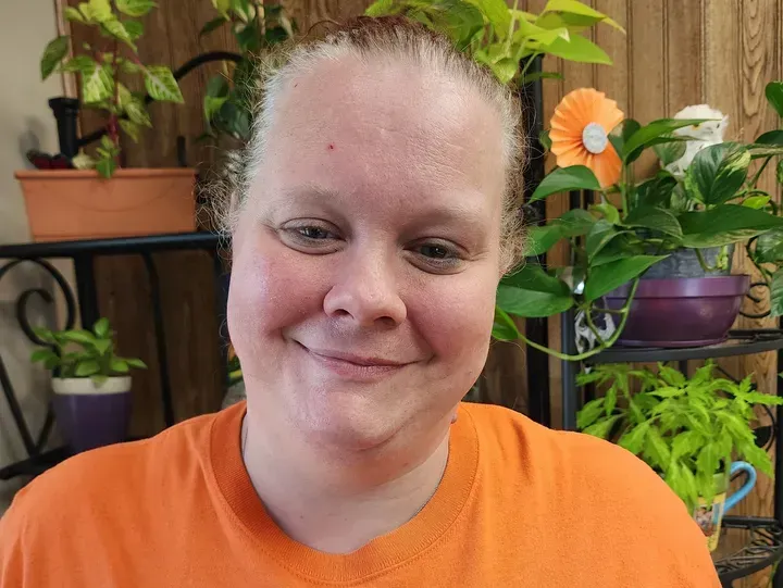 Woman in orange shirt smiles, surrounded by indoor plants on shelves.