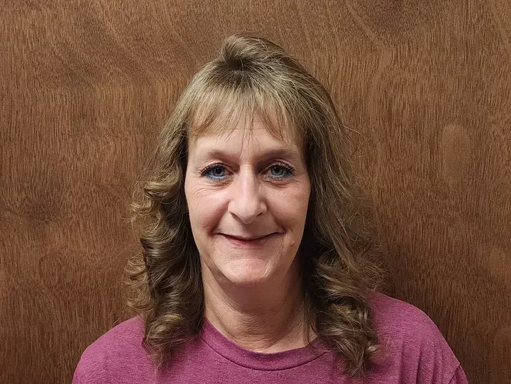 Woman with light brown hair and a pink shirt smiles in front of a wood background.