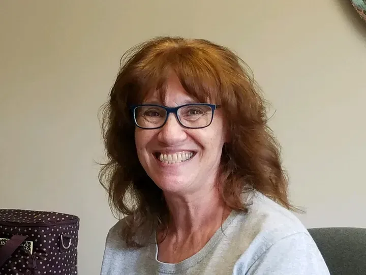 Woman with glasses smiles; brown hair, gray shirt, plain background.