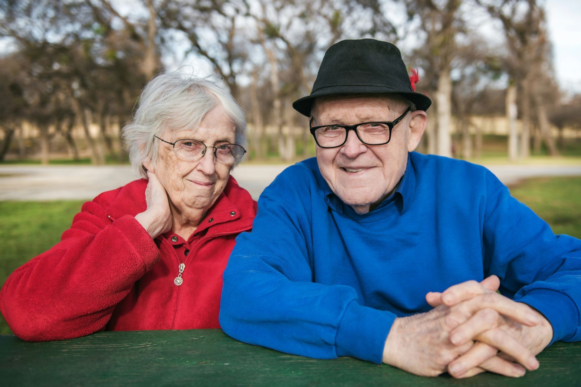 Elderly couple at a table outdoors. Woman in red jacket, man in blue sweater and fedora, both wearing glasses. Smiling.