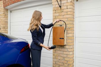 Woman charging blue electric car in front of a garage, using a wall-mounted charger.